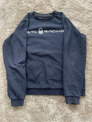 Mörkblå sweatshirt från Sail Racing - Mörkblå sweatshirt från Sail Racing med vit logotyp och text på bröstet. Tröjan har rund hals, ribbade muddar vid ärmslut och nederkant samt en liten logga på ryggen. Perfekt för en clean och sportig look. OBS! Barnstorlek 160.