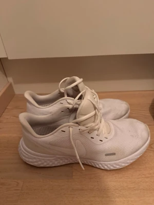Vita sneakers från Nike - Säljer ett par helvita sneakers från Nike med klassisk låg profil och snörning. Skorna har en luftig mesh-överdel och en tjock, strukturerad sula för extra komfort. Perfekta för dig som gillar stilrena och sportiga skor.