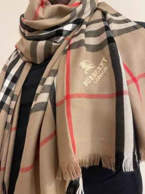 Beige rutig halsduk från Burbery - Snygg beige halsduk från Burbery med klassiskt rutmönster i svart, vitt och rött. Halsduken har fransade kanter och Burbery London-logga broderad i guld. Perfekt accessoar för att lyfta din outfit med en tidlös och lyxig känsla.