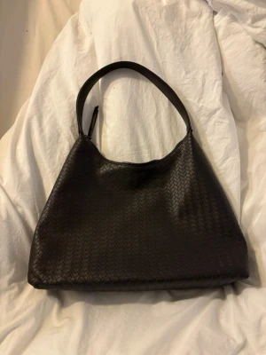 Svart flätad axelväska i skinnimitation - Snygg svart handväska med flätad struktur och mjuk, halvmåneformad design. Väskan har ett brett axelband och dragkedja upptill. Tillverkad i skinnimitation som ger en lyxig känsla. Perfekt för dig som gillar stilrena accessoarer.