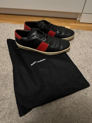 Saint Laurent svarta sneakers med röd detalj - Snygga svarta Saint Laurent sneakers i läder med röd sidopanel och vit sula. Klassisk låg modell med svarta skosnören och diskret logga på plösen. Perfekta för dig som vill ha en stilren men ändå iögonfallande sko.