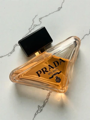 Prada Paradox Eau de Parfum - Unik trekantig parfymflaska från Prada med svart lock och guldiga detaljer. Flaskan är genomskinlig och innehåller en bärnstensfärgad doft. Stilig design med tydlig Prada-logga och texten 'Milano'. Perfekt för dig som gillar exklusiva dofter.