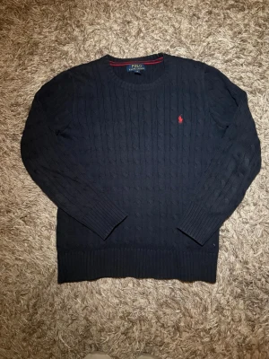 Ralph lauren stickad 14-16 år - Kabelstickad mörkblå tröja från Polo Ralph Lauren med rund halsringning och den klassiska röda logotypen broderad på bröstet. Tröjan har långa ärmar och ribbade muddar vid ärmslut och nederkant. Perfekt till sommarn och finare tillfällen. Skicket är som nytt. Hör av er vid funderingar😃