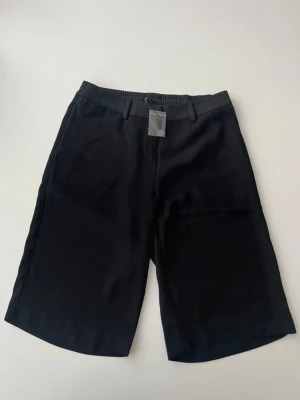 Svarta shorts från b.young - Stilrena svarta shorts från b.young med elastisk midja och hällor för bälte. Klassisk rak passform och knälång modell, perfekta för en clean och enkel look. Tillverkade i ett mjukt material som känns bekvämt hela dagen.