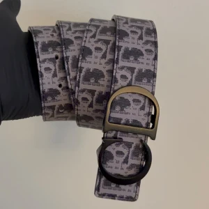 Svartvitt bälte - Snyggt grått bälte med svart döskallemönster och unik metallspänne i svart och mässing. Perfekt accessoar för att ge din outfit en edgy touch. Bältet är tillverkat i skinn och har en cool design som sticker ut.