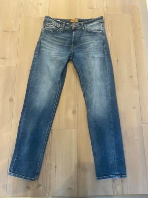 Blå raka jeans från Jack and Jones  - Säljer ett par klassiska blå jeans från Jack and Jones Je med rak passform och snyggt slitna detaljer. Jeansen har fem fickor, knappgylf och är gjorda i mjuk denim. Perfekta för en avslappnad och stilren look.