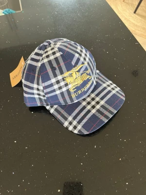 Blårutig Burberry keps - Snygg keps från Burberry i klassiskt blått rutmönster med vita och röda detaljer. Framtill finns ett gult broderat Burberry-emblem och logga. Kepsen har böjd skärm och justerbar rem bak. Tillverkad i bomull för en bekväm känsla.