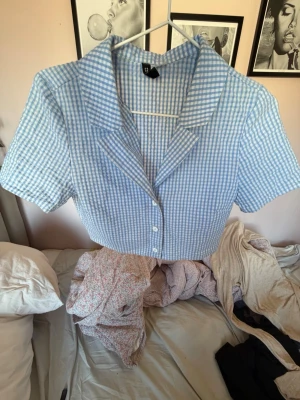 Blårutig croppad skjorta med krage - Söt croppad skjorta i blåvitt rutigt mönster med korta ärmar och klassisk krage. Skjortan har knappar framtill och en avslappnad passform. Perfekt för dig som gillar retrovibbar och vill ha något lätt till sommaren.