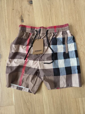 Rutig Burberry shorts med snörning - Säljer ett par klassiska Burberry shorts med det ikoniska rutiga mönstret i beige, svart, vitt och rött. Shortsen har snörning i midjan och sidofickor. Perfekta för en trendig och avslappnad stil.