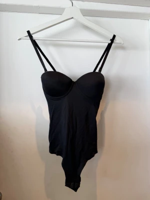 Svart body med bygel och smala band - Snygg svart body med bygelkupor och justerbara smala axelband. Bodyn har knäppning i grenen med tryckknappar för enkel på- och avtagning. Materialet är stretchigt och följsamt, perfekt för en stilren look. 