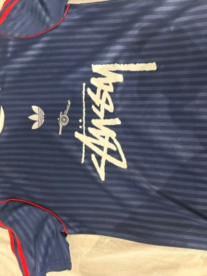 Blå randig Stüssy x Adidas t-shirt - Säljer en blå randig t-shirt från Adidas i samarbete med Stüssy. T-shirten har vit Stüssy-logga framtill, Adidas- och Arsenal-detaljer samt röda ränder på ärmarna. Slim fit modell i syntetmaterial, perfekt för dig som gillar streetwear.