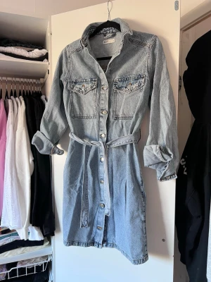 Jeansklänning med knytskärp och knappar, stl 36 - Superfin jeansklänning i ljusblå denim med långa ärmar, knappar hela vägen framtill och två bröstfickor. Knytband i midjan för en markerad siluett. Perfekt nu till vår/sommar!!