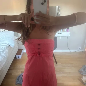 Axelbandslös korall klänning - Så sommrig och fin färg. Snygga knappar. Har resor band bak till och därför väldigt stretchig. Snygga knappar. Köpt på Plick men aldrig använt och har inga defekter