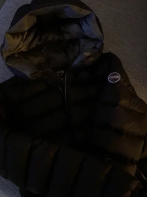 Svart jacka från Colmar - Snygg svart pufferjacka från Colmar med huva och dragkedja framtill. Jackan har ett quiltat mönster och Colmar-logga på bröstet. Perfekt för kalla dagar och ger en trendig look. Passar till det mesta och är riktigt skön att bära.