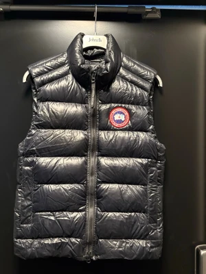 Svart dunväst från Canada Goose - Snygg svart dunväst från Canada Goose med hög krage och dragkedja framtill. Västen är perfekt inför våren och även sommaren, äkta från Johnells i Örebro