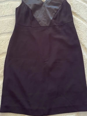 Svart klänning från Yessica - Stilren svart klänning från Yessica med v-ringad överdel och figurnära passform. Klänningen har en klassisk siluett och är tillverkad i ett mjukt tyg som känns bekvämt mot huden. Perfekt för dig som gillar enkel men elegant stil.