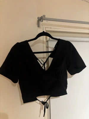 Svart croppad topp med knytband - Trendig svart croppad topp med djup v-ringning och öppen rygg. Toppen har snygga knytband bak som ger en cool detalj och justerbar passform. 