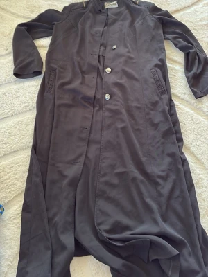 Lång mörkgrå kappa från Elite Defile - Snygg lång mörkgrå kappa från Elite Defile med knappar framtill och diskreta fickor med dekorativa detaljer. Tillverkad i Turkiet. Perfekt för dig som vill ha en stilren och enkel look med en touch av elegans.