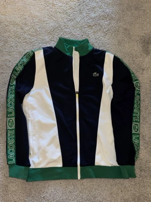 Lacoste sportjacka blå/vit/grön - Sportig jacka från Lacoste i blått, vitt och grönt med hel dragkedja och hög krage. LACOSTE-logga på bröstet och längs ärmarna. Tillverkad i mjukt material och har gröna muddar vid ärmslut och nederkant.