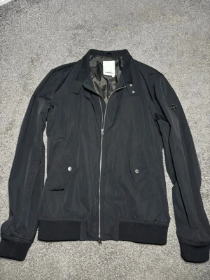 Svart vindjacka från J.Lindeberg - Stilren svart vindjacka från J.Lindeberg med silverfärgad dragkedja, två snedställda fickor med tryckknappar och ribbade muddar vid ärmslut och nederkant. Jackan har en diskret logotyp på ärmen och är tillverkad i ett lätt, vindtätt material.