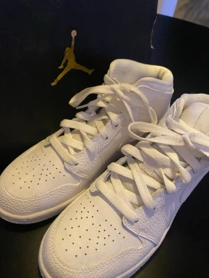 Vita Air Jordan 1 Mid sneakers barn - Säljer ett par vita Air Jordan 1 Mid sneakers med klassisk siluett, perforerad tåbox och snörning. Skorna är tillverkade i skinn och har en stilren look med högt skaft. Perfekta för dig som gillar ikoniska sneakers och vill ha något som sticker ut. Storlek: eur 36.5 