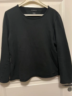 Svart långärmad tröja från Tiger of Sweden - En stilren svart långärmad tröja från T/J med rund halsringning. Tröjan är tillverkad i mjukt bomullsmaterial och har en diskret logga nertill. Perfekt för en enkel och clean look.