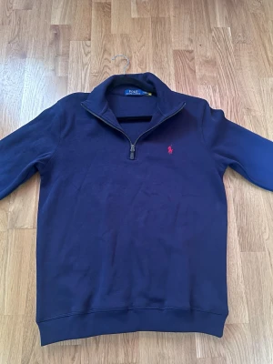 Ralph Lauren tröja - Mörkblå stickad tröja från Polo Ralph Lauren med halv dragkedja och klassisk röd logga broderad på bröstet. Tröjan har hög krage och ribbade muddar vid ärmslut och nederkant. Perfekt för lager på lager och stilren look.