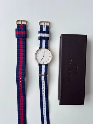  Äkta Daniel Wellington 36mm Guld – 2 Tygband (Vitt/Marin + Rött/Marin) - Snygg Daniel Wellington-klocka 36 mm i guld, sparsamt använd och i nästan nytt skick.  Originalförpackning finns kvar till extra-bandet men inte till själva klockan.
