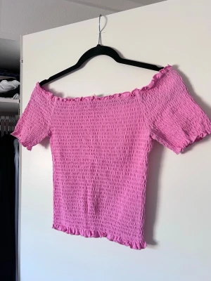 Rosa offshoulder topp, stl M - Söt rosa offshoulder topp med korta ärmar och volangdetaljer vid kanterna. Toppen har en smockad, stretchig struktur som ger en tight passform.