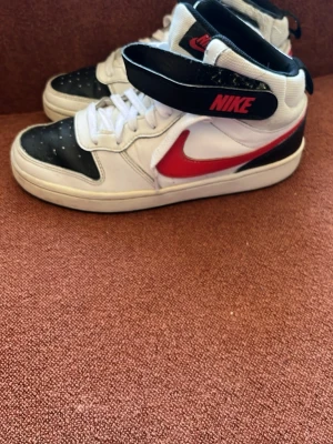 Nike vita och svarta high sneakers - Nike sneakers i high-modell med vit bas, svarta detaljer på tån och runt ankeln samt röd swoosh och Nike-logga på kardborrebandet. Skorna har klassisk snörning och perforerade partier framtill. Tillverkade i syntetmaterial och textil.