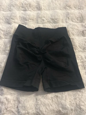 Svarta shaping shorts - Svarta shaping shorts med hög midja och sömlös design. Materialet är stretchigt och formar kroppen snyggt. Perfekta under klänningar eller kjolar för en slät siluett. Shortsmodellen har diskret rynk bak för extra passform.