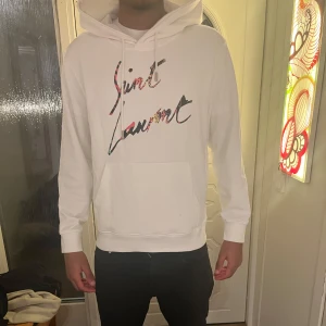Sällsynt Saint Laurent hoodie  - Säljer en stilren vit hoodie från Saint Laurent med färgglad logotyp på bröstet. Tröjan har en klassisk känguruficka och justerbar huva med snören. Perfekt för en avslappnad look. Modellen är 180 och väger cirka 65 