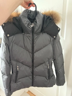 Grå Moncler dunjacka med pälskrage - Snygg grå dunjacka från Moncler med stor huva och avtagbar pälskant i brunt. Jackan har två dragkedjefickor fram, hög krage med spänne och tydlig Moncler-logga på ryggen. Puffig modell som håller dig varm under kalla dagar.