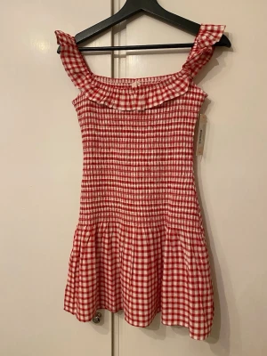 Rödvit rutig offshoulder klänning - Supersöt kortklänning i rödvitt rutigt mönster med offshoulder och volangkant upptill. Smockad överdel ger en tight passform och kjolen faller lätt. Perfekt för sommarens alla tillfällen. Materialet känns som bomull och klänningen har en ungdomlig vibe.