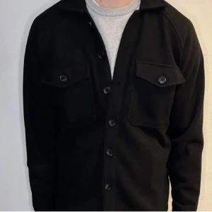 Svart overshirt med fickor - Stilren svart overshirt med knäppning framtill och två stora bröstfickor med lock och knapp. Klassisk krage och raka ärmar. Perfekt lager-på-lager-plagg för en avslappnad look. Materialet ser ut att vara mjukt och något tjockare, vilket ger en schysst passform.
