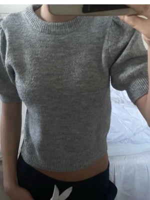 Grå stickad topp med puffärm - Säljer en grå stickad topp med korta puffärmar och ribbade muddar vid hals, ärmslut och nederkant. Tröjan har en croppad passform och är gjord i mjukt material som känns skönt mot huden. Perfekt för dig som gillar en enkel men trendig look.
