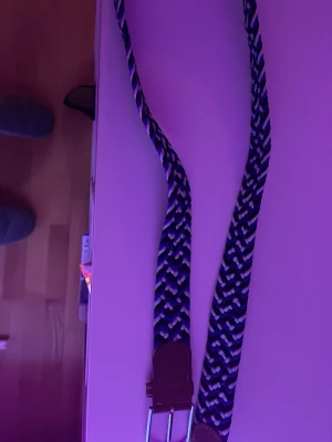 Blåvitt flätat bälte med läderdetalj - Snyggt flätat bälte i blått, vitt och svart med silverfärgat spänne och bruna läderdetaljer vid spännet. Perfekt accessoar för att lyfta din outfit med en sportig och fräsch känsla.