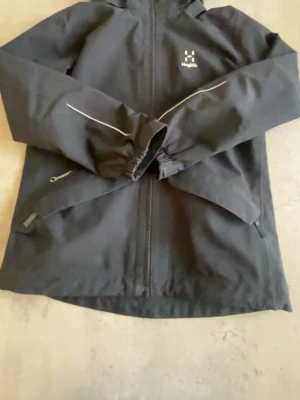 Svart regnjacka från Haglöfs - Pris går att diskutera Svart regnjacka från Haglöfs med huva och hel dragkedja framtill. Jackan har reflexdetaljer på ärmarna och två snedställda fickor med dragkedja. Tillverkad i slitstarkt, vattenavvisande material. Perfekt för regniga dagar.