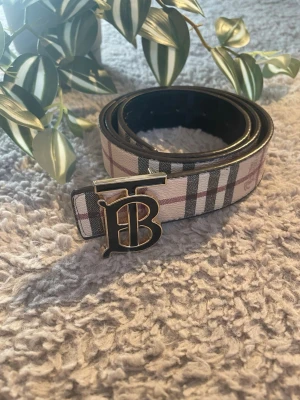 Bälte - Snyggt bälte med klassiskt rutmönster i beige, svart och rött. Bältet har ett stort metallspänne med TB-logga framtill och är gjort i läder med tygdetaljer. Perfekt accessoar för att lyfta vilken outfit som helst.