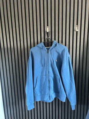 Blå Jordan zip hoodie  - Blå zip hoodie från Jordan med huva och dragsko. Hoodien har två stora fickor framtill och en liten broderad Jumpman-logga på bröstet. Tillverkad i mjukt sweatshirtmaterial, perfekt för en avslappnad och sportig stil.