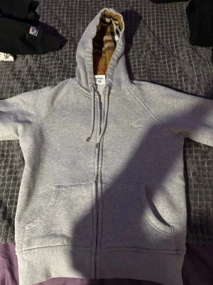 Grå zip hoodie från Burberry - Snygg grå zip hoodie från Burberry med klassiskt rutigt foder i huvan och dragsko. Hoodien har två stora fickor framtill och diskret Burberry-logga på bröstet. Perfekt för dig som vill ha en stilren och exklusiv look.