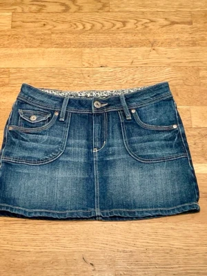 Kort jeanskjol med fickor H&M - Snygg blå kort jeanskjol med klassiska fickor fram och bälteshällor från H&M. Kjolen har en knapp och dragkedja framtill samt en fin blommig kant på insidan av linningen. Perfekt för en trendig och avslappnad stil. Storlek 152, men passar mig som är XS.