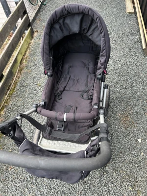 Svart barnvagn med regnskydd - Säljer en svart barnvagn med justerbart handtag, regnskydd och säkerhetssele. Vagnen har en robust metallram och vadderad sits för extra komfort. Passar bra för promenader och har praktisk förvaringskorg under sitsen.