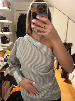 Randig off shoulder blus - Säljer en snygg off shoulder blus med gråvita vertikala ränder från Zara.  Blusen har en unik asymmetrisk design med en ärm och knappar i sidan. Tillverkad i ett mjukt bomullsliknande material som känns bekvämt mot huden. Öppen detalj i ryggen 