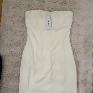 Vit axelbandslös kort klänning - Säljer en stilren vit axelbandslös klänning från NA-KD. Klänningen är figurnära och kort den hamnar ungeför lite över knäna. Aldrig användt innan därav är etiketten kvar.