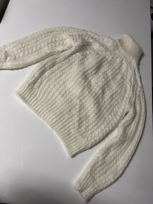 Vit kabelstickad polotröja - Kabelstickad polotröja i mjukt vitt garn med hög krage och långa ärmar. Tröjan har ribbade muddar vid ärmslut och nederkant samt ett klassiskt mönster som ger en mysig känsla. Perfekt för kyliga dagar och enkel att styla till olika outfits.