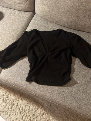 Svart v-ringad blus med puffärm - Svart blus med v-ringning och trekvarts puffärmar. Materialet är ribbat och ger en snygg struktur. Passformen är normal och blusen är lätt att matcha med olika outfits.