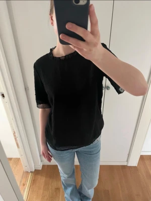 Svart blus med spetsdetaljer - Säljer en svart blus med halvtransparent tyg och fina spetsdetaljer vid halsringning och ärmslut. Blusen har korta ärmar och en lös passform som ger en avslappnad men ändå stilren look. Perfekt att matcha med jeans eller kjol.