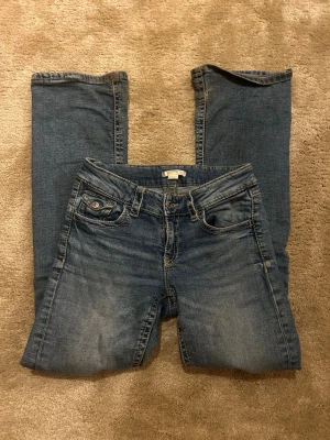 Blå bootcut jeans med fickdetaljer - Snygga blå bootcut jeans med klassisk femficksdesign och coola ficklock med knappar bak. Jeansen har en lätt tvättad look och är gjorda i denim med lite stretch för skön passform. Perfekta till sneakers eller boots.