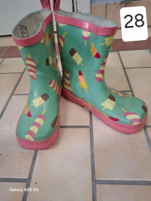 Gröna gummistövlar med glassmotiv - Färgglada gummistövlar i grönt med rosa kant och sula. Stövlarna har ett lekfullt mönster av olika glassar i flera färger. Perfekta för regniga dagar och för dig som gillar roliga detaljer på skorna.
